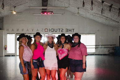 Five women wearing cowgirl hats, pink western outfits and bandanas pose smiling in a mirrored dance studio with hardwood floor and ceiling fans.