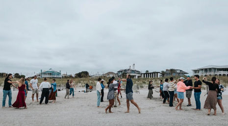 Couples and friends dancing barefoot on a sandy beach near dunes and coastal houses under an overcast sky
