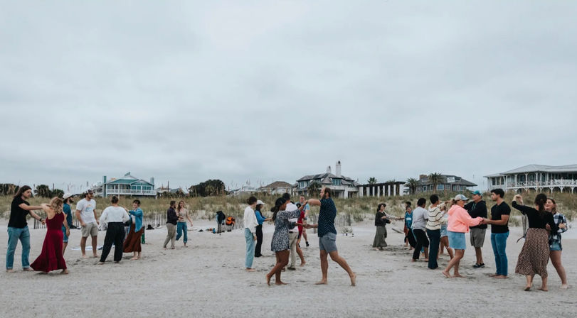 Couples and friends dancing barefoot on a sandy beach near dunes and coastal houses under an overcast sky