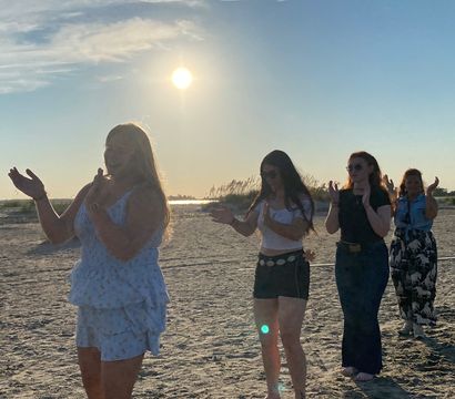Group of people clapping in a line on a sandy beach at golden-hour sunset, sun low over coastal grasses and shimmering water, casual summer outfits and relaxed atmosphere.