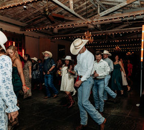 Guests line-dancing at a rustic indoor barn reception, wearing cowboy hats and boots beneath string lights and chandeliers — lively country wedding scene.