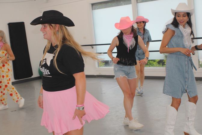 Group of women line dancing in a bright indoor studio wearing cowboy hats and western-inspired outfits — pink skirt, denim dress, shorts and white boots.