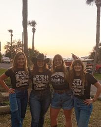 Four smiling friends in denim and black graphic tees—one wearing a cowboy hat—posing arm-in-arm at a palm‑tree lined coastal sunset with parked cars and beach gear in the background.