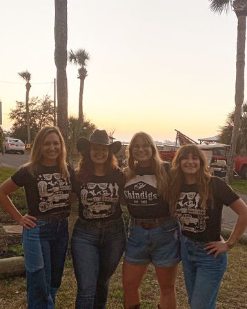 Four smiling friends in denim and black graphic tees—one wearing a cowboy hat—posing arm-in-arm at a palm‑tree lined coastal sunset with parked cars and beach gear in the background.