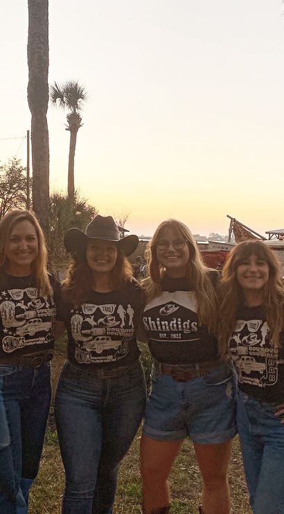 Four smiling friends in black graphic tees and denim pose arm‑in‑arm at a coastal sunset framed by tall palm trees — one wears a cowgirl hat and another glasses, with parked vehicles and a shoreline silhouette behind them.