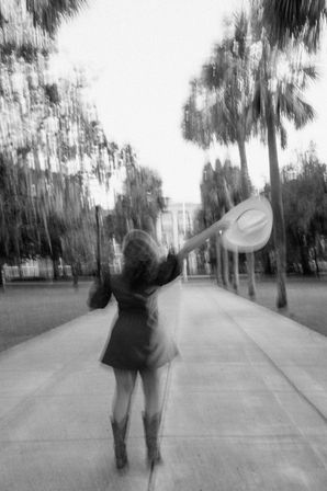 Black-and-white motion-blur photo of a person walking away down a palm-lined walkway, arm raised waving a wide-brim hat, wearing a short dress and cowboy boots — carefree outdoor scene.