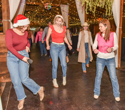 Group line dancing in a rustic barn venue under twinkling string lights, women in jeans and boots with a Santa hat and cowboy hat at a festive holiday party.