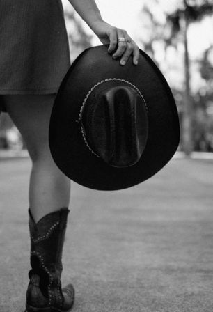 Black-and-white photo of a person in cowboy boots and a short dress, casually holding a wide-brimmed cowboy hat by their side on a blurred street.