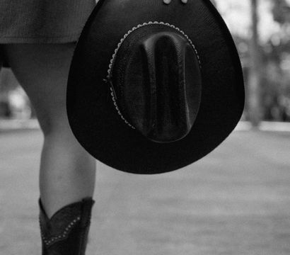Black-and-white photo of a person in cowboy boots and a short dress, casually holding a wide-brimmed cowboy hat by their side on a blurred street.