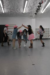 Six women in a bright dance studio wearing cowboy hats, boots and pink accents, creating a playful high-five arch as others step through during a country-style dance rehearsal.