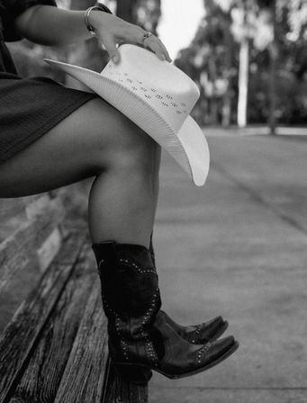 Black-and-white photo of a seated figure on a wooden bench holding a white cowboy hat, legs crossed and wearing studded cowboy boots beside a tree-lined sidewalk.