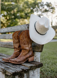 Worn brown leather cowboy boots and a white cowboy hat resting on a weathered wooden bench in a sunlit country yard.