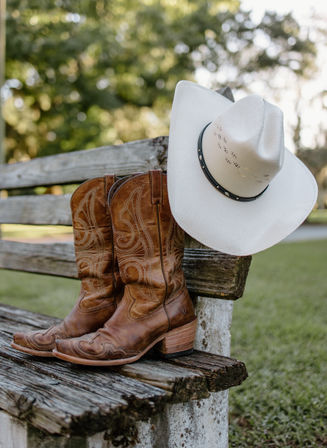 Worn brown leather cowboy boots and a white cowboy hat resting on a weathered wooden bench in a sunlit country yard.
