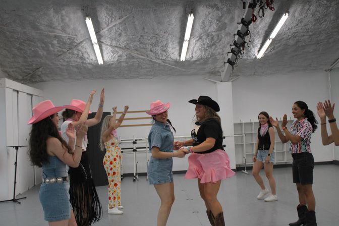 Group of women in an indoor dance studio wearing pink and black cowboy hats and western outfits, laughing, clapping and dancing during a country-themed rehearsal.