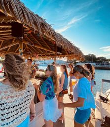 Sunlit group at a thatched-roof tiki bar on a marina dock, sipping drinks and chatting in casual summer clothes during golden-hour.