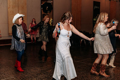 Group of women dancing on an indoor dance floor at a country-themed party — woman in a white dress holding a beer bottle leads while others wear cowboy boots, sequined dresses and a white cowboy hat.
