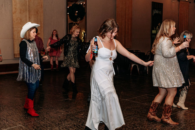 Group of women dancing on an indoor dance floor at a country-themed party — woman in a white dress holding a beer bottle leads while others wear cowboy boots, sequined dresses and a white cowboy hat.