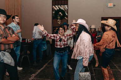 Group of adults dancing at an indoor western-themed party — cowboy hats, fringed jackets, plaid shirts and boots on a rustic dance floor.