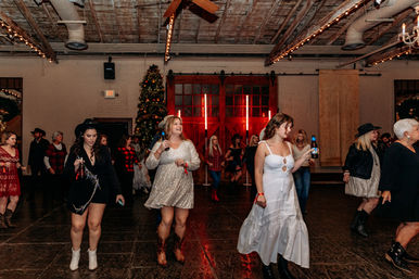 Women dancing on a rustic indoor dance floor at a holiday party — string lights, Christmas tree, red barn doors, cowboy boots, cocktails and festive attire.