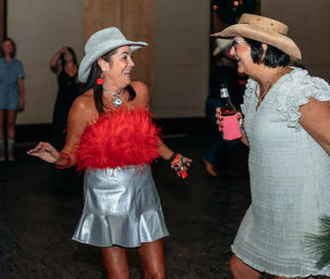 Two women in cowboy hats laughing and dancing on an indoor party dance floor — one in a silver skirt and red feather top, the other in a light ruffled dress holding a beer bottle with a pink koozie.