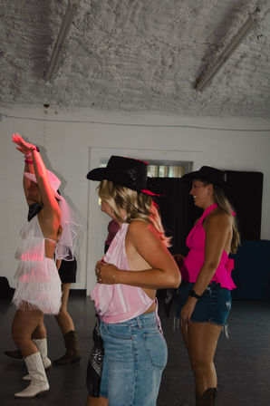 Group of friends line-dancing at a Western-themed bachelorette party indoors — bride in a white veil and fringe dress, others in cowboy hats, denim, cowgirl boots and bright pink tops.