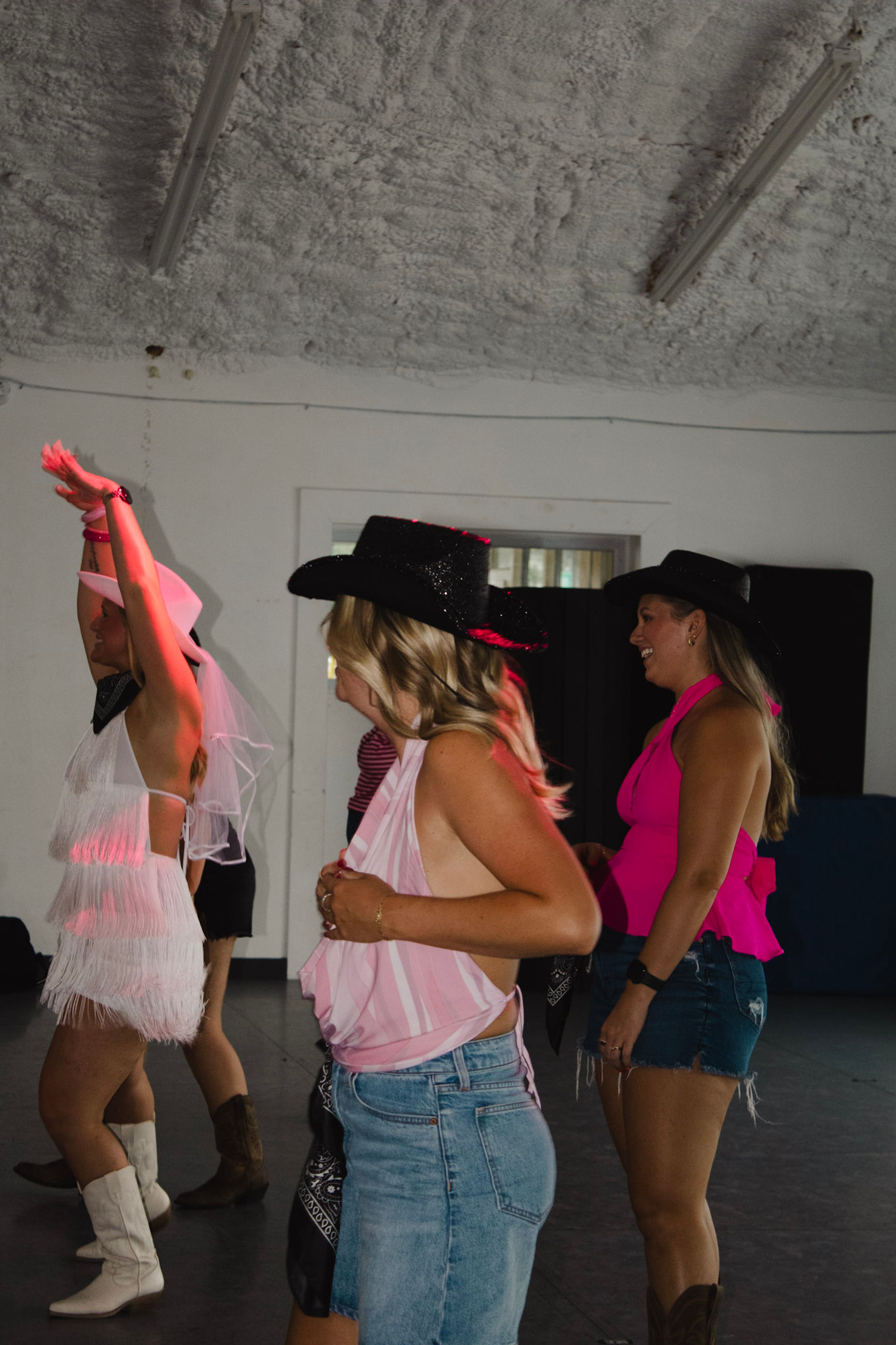 Group of friends line-dancing at a Western-themed bachelorette party indoors — bride in a white veil and fringe dress, others in cowboy hats, denim, cowgirl boots and bright pink tops.