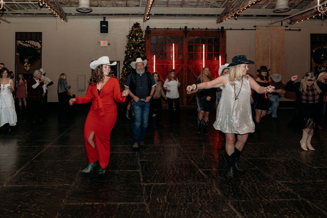Group line-dancing at a country-western holiday party in a rustic indoor venue with barn-style red doors and a decorated Christmas tree, people wearing cowboy hats and festive dresses on a dark dance floor.