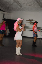 Women line-dancing in an indoor studio in cowgirl outfits — center dancer in a white fringe dress, pink veiled cowboy hat and white boots, with a "Welcome Back 2026" sign on the wall.