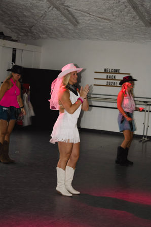 Women line-dancing in an indoor studio in cowgirl outfits — center dancer in a white fringe dress, pink veiled cowboy hat and white boots, with a "Welcome Back 2026" sign on the wall.