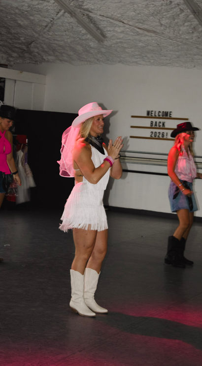 Women line-dancing in an indoor studio in cowgirl outfits — center dancer in a white fringe dress, pink veiled cowboy hat and white boots, with a "Welcome Back 2026" sign on the wall.