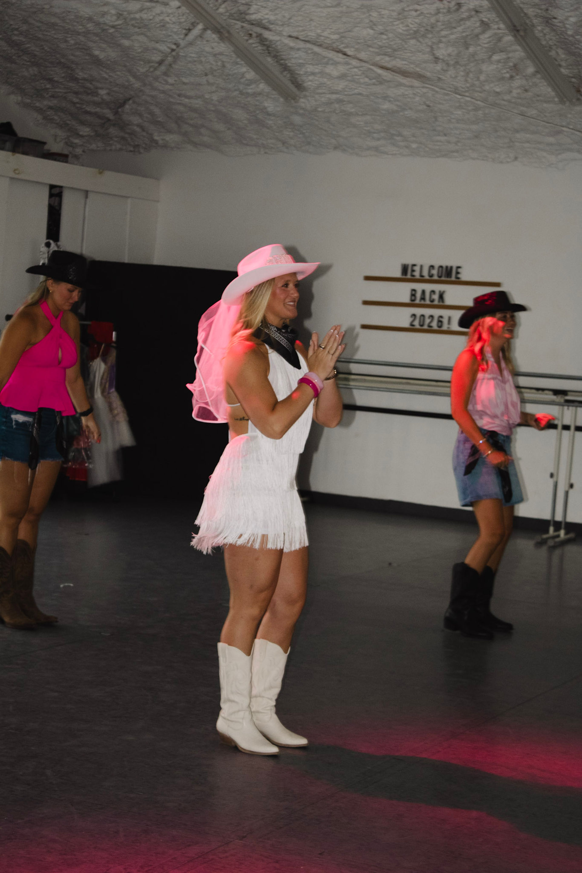 Women line-dancing in an indoor studio in cowgirl outfits — center dancer in a white fringe dress, pink veiled cowboy hat and white boots, with a "Welcome Back 2026" sign on the wall.