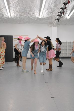 Group of women in denim outfits, pink cowboy hats and boots laughing through a high-five tunnel in a bright indoor dance studio