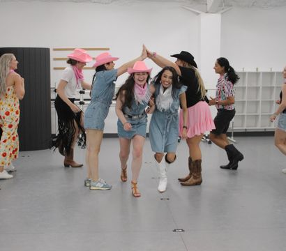 Group of women in denim outfits, pink cowboy hats and boots laughing through a high-five tunnel in a bright indoor dance studio