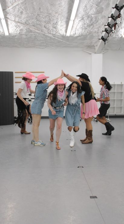Group of women in denim outfits, pink cowboy hats and boots laughing through a high-five tunnel in a bright indoor dance studio