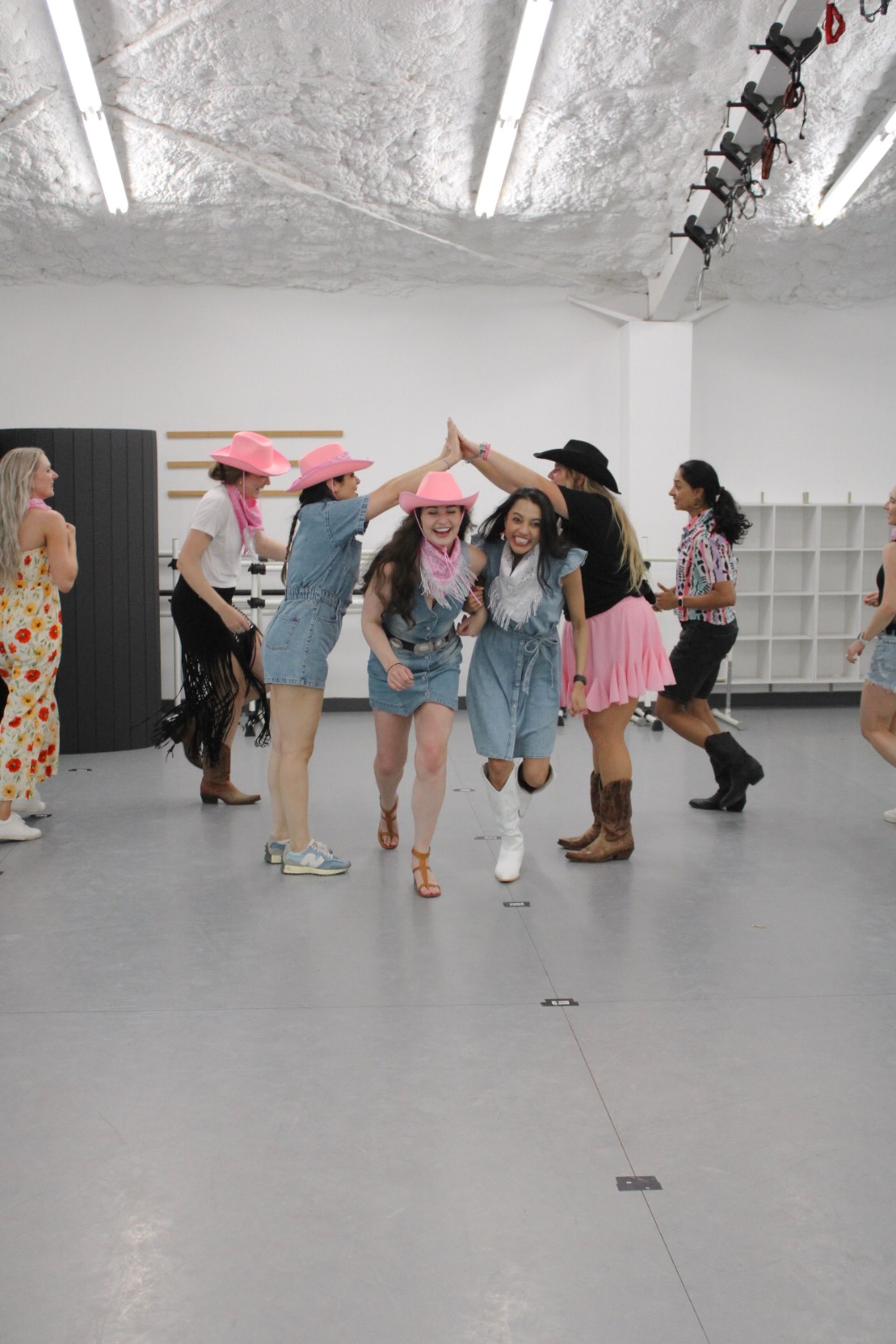 Group of women in denim outfits, pink cowboy hats and boots laughing through a high-five tunnel in a bright indoor dance studio