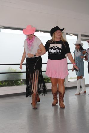 Two women line-dancing indoors on a studio floor in western attire — one in a pink cowboy hat, pink bandana and black fringed skirt, the other in a black cowboy hat, pink skirt and cowboy boots, smiling.