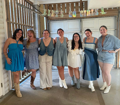 Seven friends in summer dresses and denim smiling under a 'LET'S GO GIRLS' banner in a rustic covered garage, many wearing cowboy boots, casual celebration group photo.