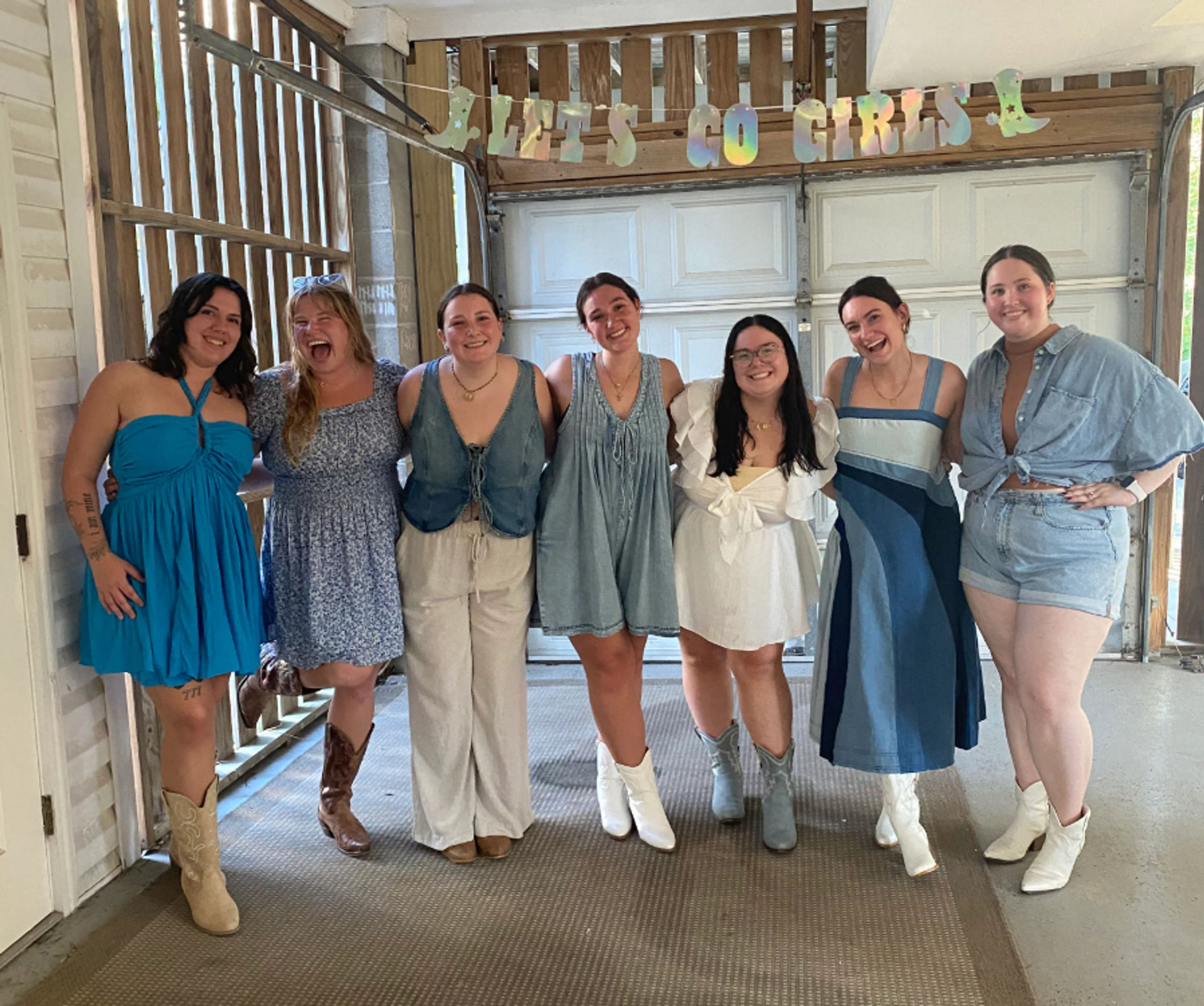 Seven friends in summer dresses and denim smiling under a 'LET'S GO GIRLS' banner in a rustic covered garage, many wearing cowboy boots, casual celebration group photo.