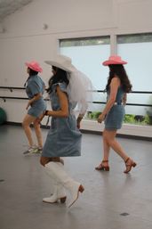 Three women in a bright dance studio doing a cowgirl line dance in denim dresses and cowboy hats — one wearing a white hat with a veil and white boots, two in pink hats and casual shoes.