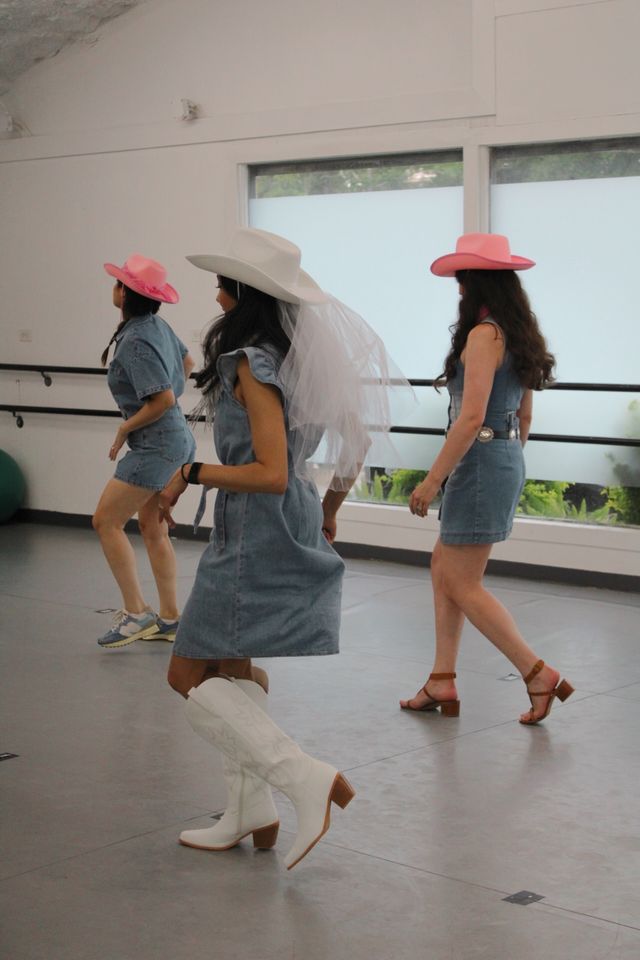 Three women in a bright dance studio doing a cowgirl line dance in denim dresses and cowboy hats — one wearing a white hat with a veil and white boots, two in pink hats and casual shoes.
