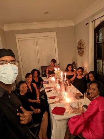 Candlelit indoor dinner party in a home dining room — a masked host takes a selfie with a group of guests seated around a table set with candles, glassware and sparkly placemats, dressed up for an intimate evening celebration.