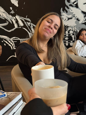 Smiling person reaching a latte across a table in a cozy coffee shop with a black-and-white mural and a friend seated in the background.