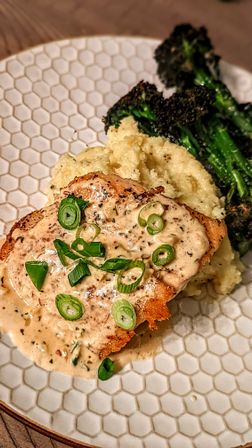 Seared chicken breast smothered in creamy herb sauce and sliced scallions, plated with mashed cauliflower and charred broccolini on a white hex-pattern dinner plate — cozy comfort dinner.
