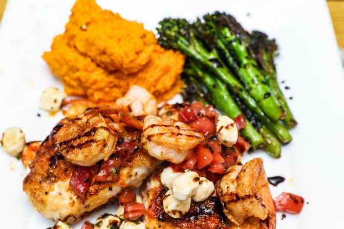 Grilled shrimp and seasoned chicken topped with diced tomato and mozzarella pearls, served with charred broccolini and creamy sweet potato mash on a white plate.