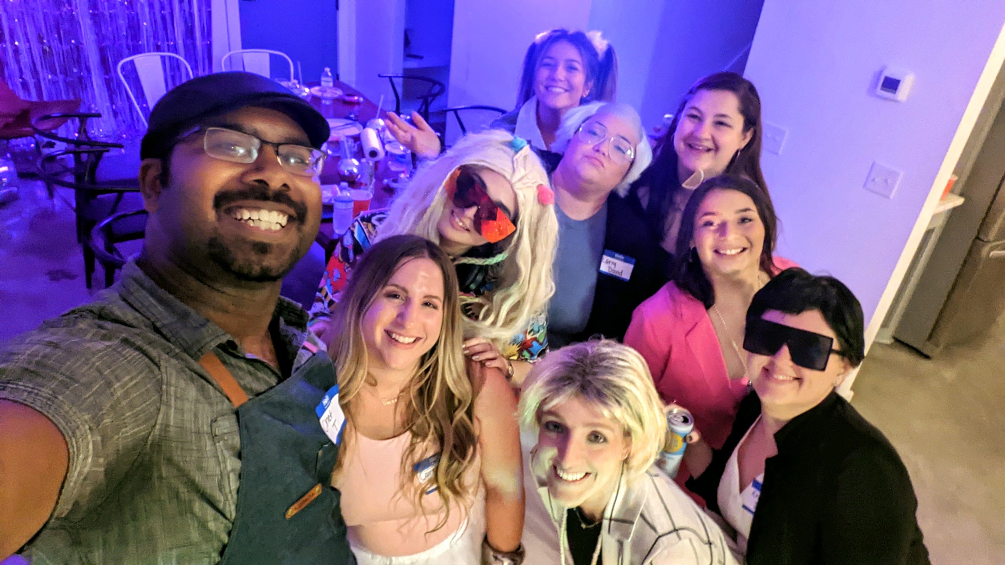 Nine friends smiling in a selfie at a colorful indoor apartment party with purple-blue lighting, drinks on tables, playful costumes, wigs and oversized sunglasses.