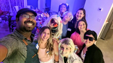 Nine friends smiling in a selfie at a colorful indoor apartment party with purple-blue lighting, drinks on tables, playful costumes, wigs and oversized sunglasses.