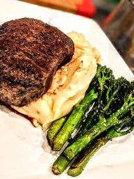 Close-up of a juicy seared filet steak on creamy mashed potatoes with charred roasted broccolini on a white plate, classic steak dinner