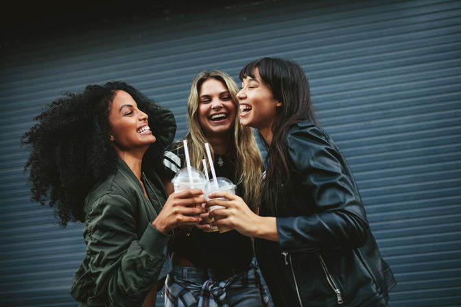 Three women laughing and clinking iced drinks in plastic cups with straws against a gray corrugated metal wall — casual urban street vibe.