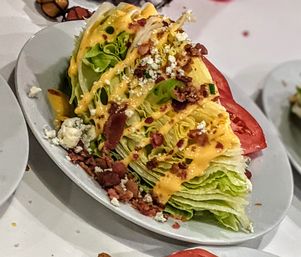 Restaurant-style wedge salad featuring a crisp iceberg lettuce wedge topped with crumbled bacon, blue cheese, tomato slices and a creamy cheddar-style dressing drizzle on a white plate