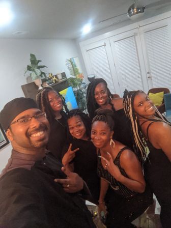 Group selfie of seven friends smiling in a cozy living room — indoor home gathering with plants, colorful pillows, closed blinds and a ceiling fan.